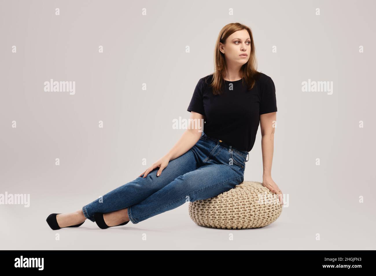 Die junge Frau in legerem schwarzen T-Shirt und Jeans sitzt auf dem Studioboden auf grauem Hintergrund Stockfoto