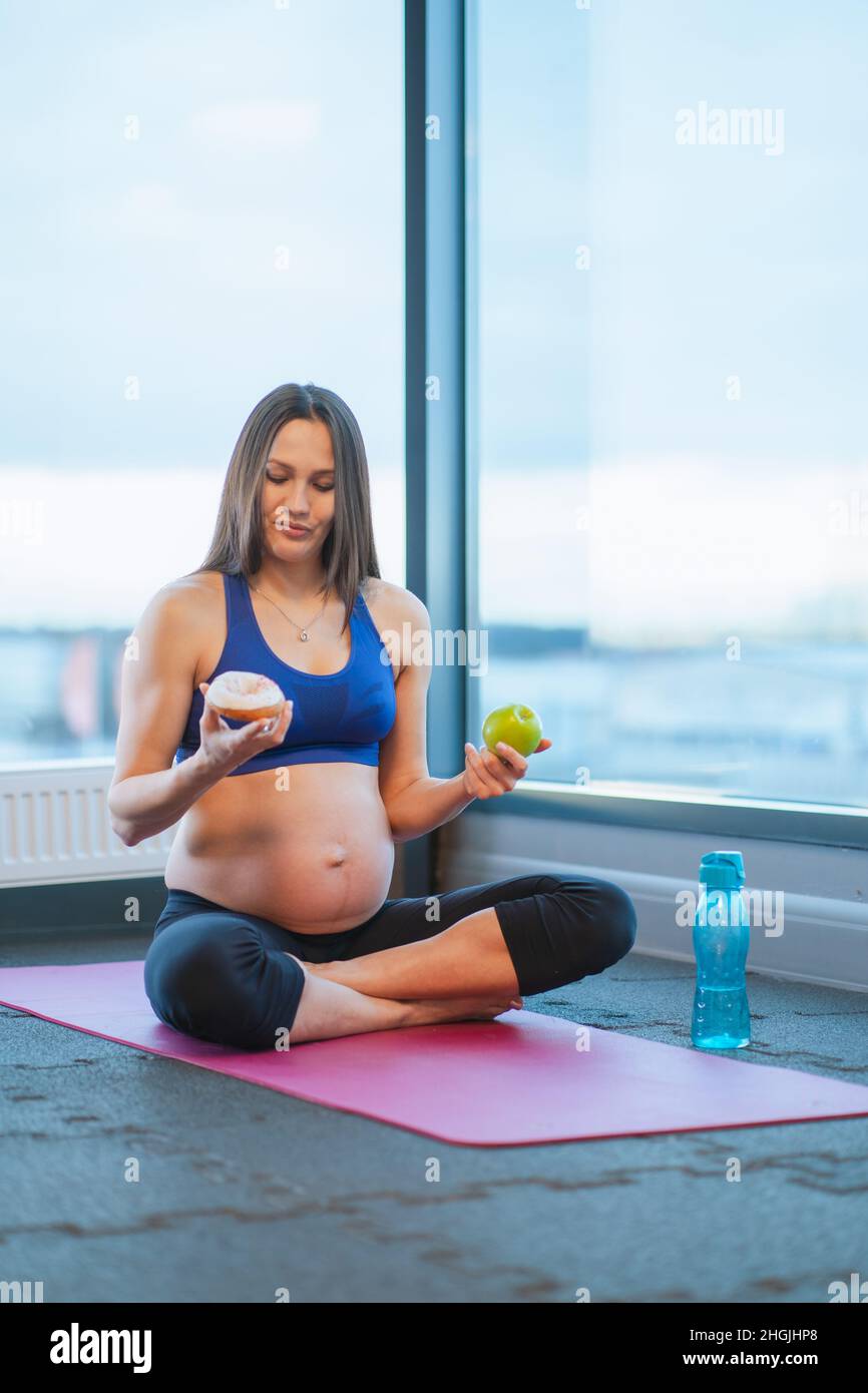 Athletische Schwangere in Sportkleidung halten Donut und Apfel auf Yoga-Matte sitzen Stockfoto