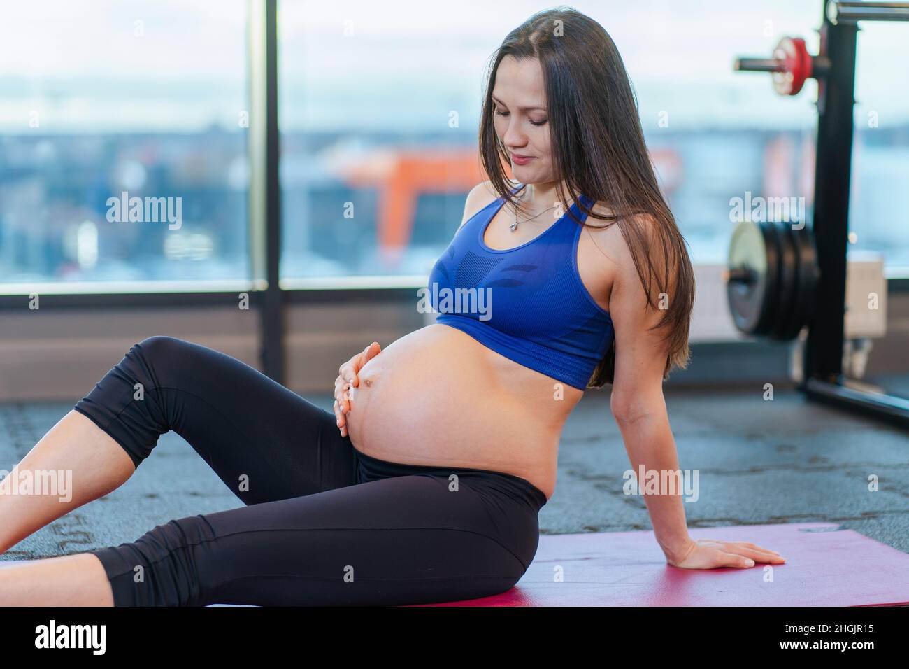 Schwangere mit langen Haaren in Sportswear ruhen sich aus und sitzen im Fitnessstudio auf einer rosa Matte. Stockfoto