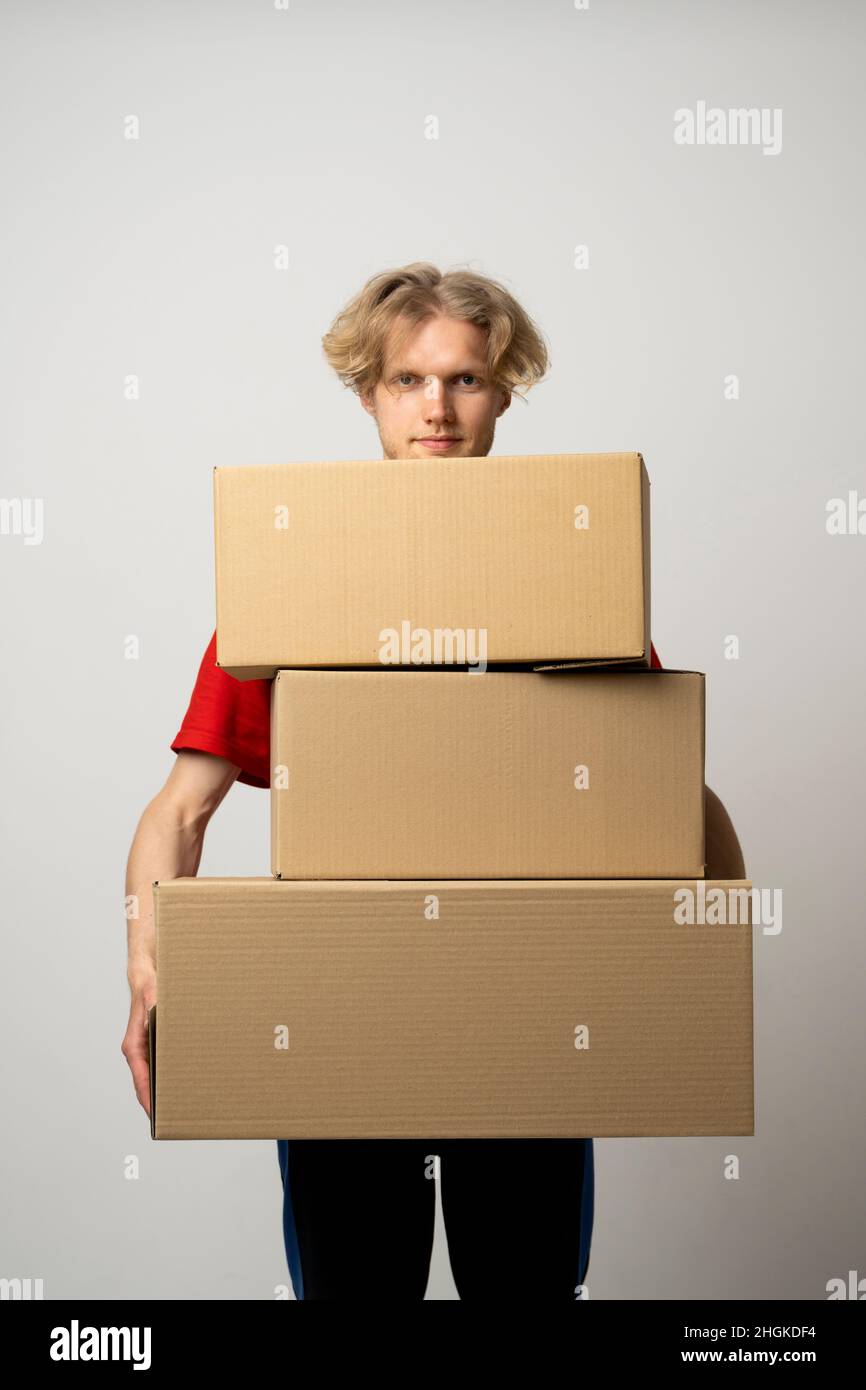 Fröhlicher Lieferer. Fröhlicher junger Kurier, der eine Pappschachtel hält und lächelt, während er vor weißem Hintergrund steht. Der Lieferer hält einen Haufen Stockfoto