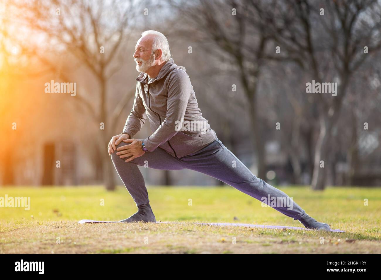 Älterer Mann mit Bart in Sportkleidung, der im Spätherbst bei Sonnenuntergang in der Natur trainiert und seine Beine streckt Stockfoto