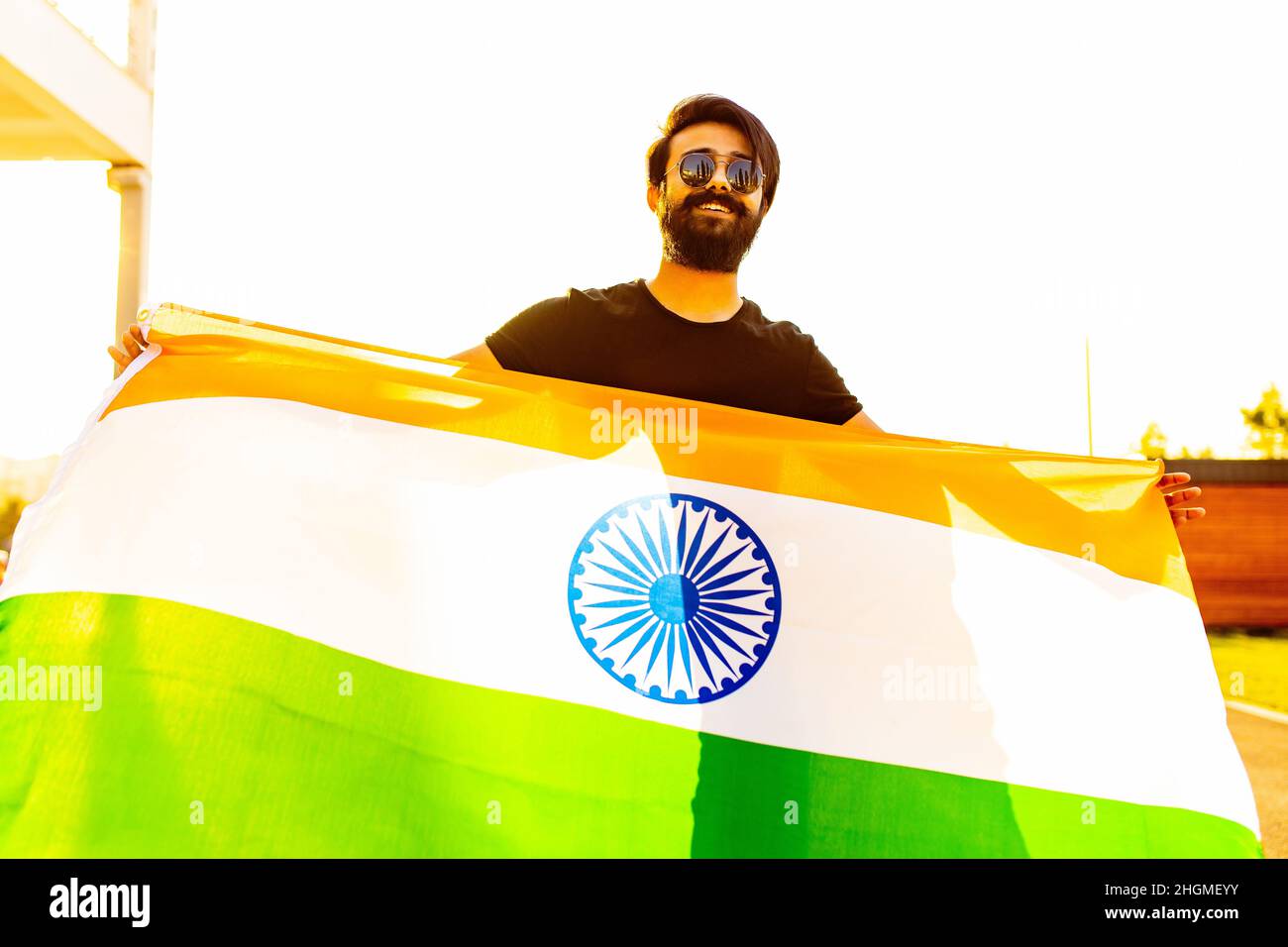 Hübscher indischer bärtiger Mann in schwarzem Baumwoll-T-Shirt, der im Sommerpark bei Sonnenuntergang bei Golglicht die indische Flagge weckte Stockfoto