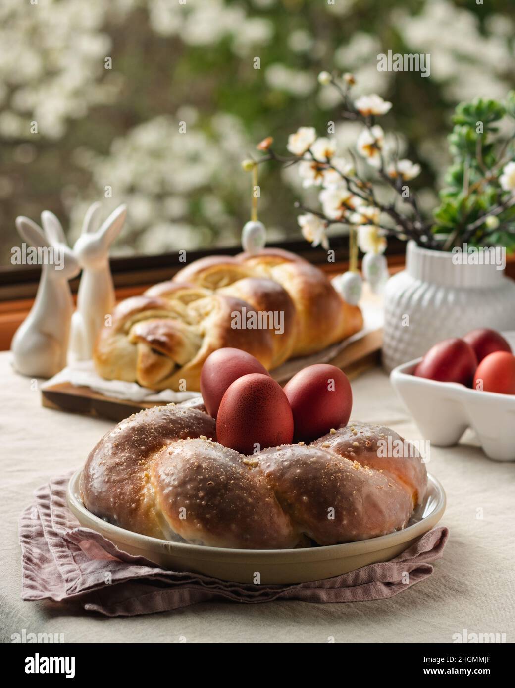 Osterbrot, österreichischer osternzopf, griechischer Tsoureki und rote Eier auf einem Tisch mit Leinentischdecke mit Blick auf das Frühlingsfenster, Stillleben Stockfoto