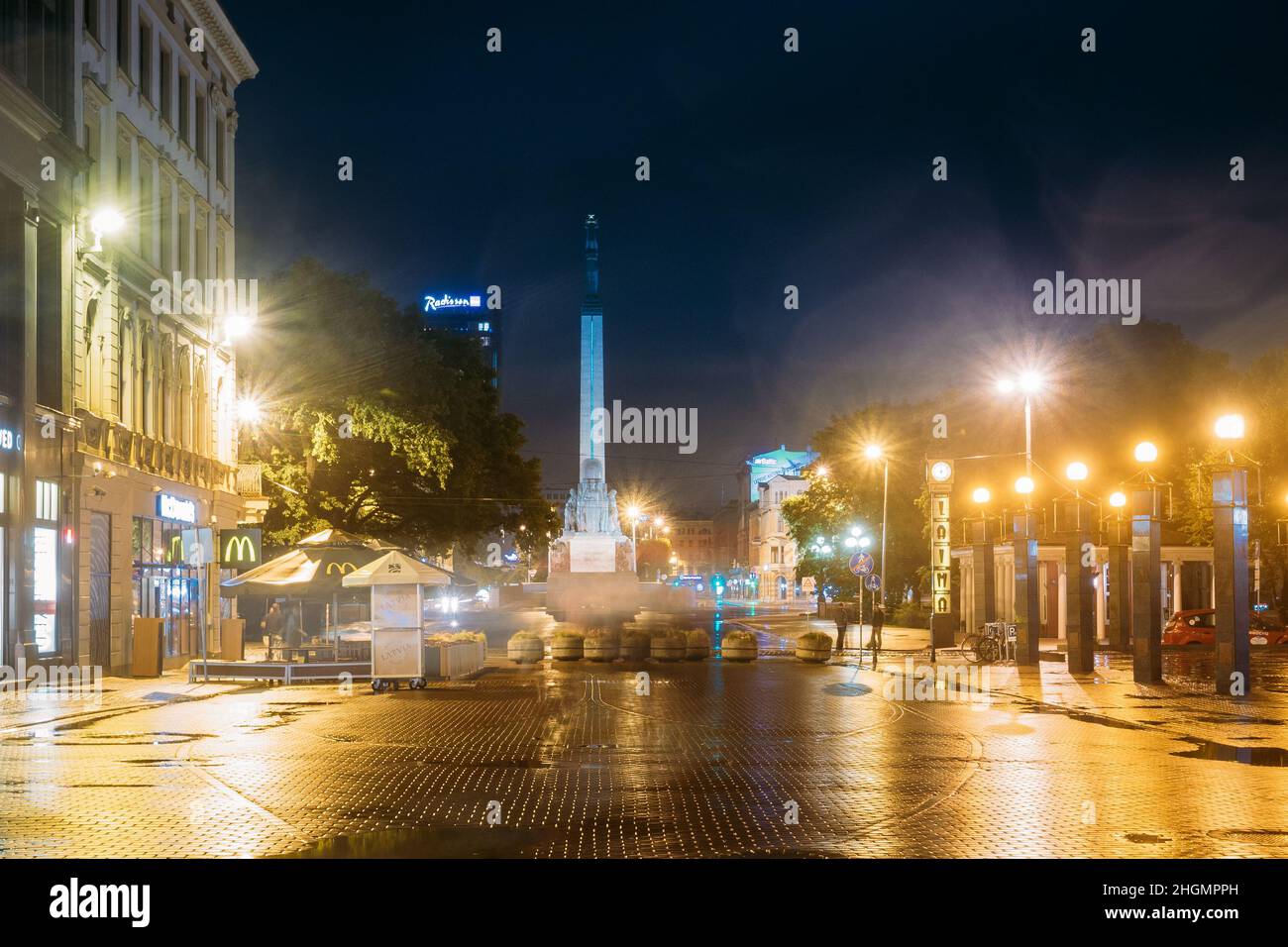 Riga, Lettland. Nachtansicht Des Memorial Freedom Monument Am Freedom Square Und Laima Clock Lighting Bei Nachtbeleuchtung. Blick Von Der Kalku Street. Stockfoto