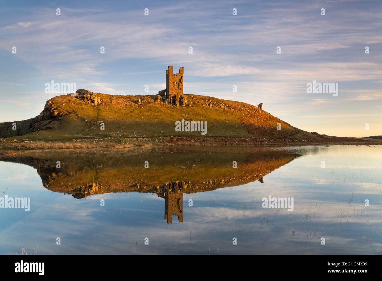 Die Ruinen von Lilburn Tower, Dunstanburgh Castle an der Küste von Northumberland, spiegeln sich in den ruhigen stillen Gewässern eines Sees bei Sonnenuntergang im Winter wider Stockfoto