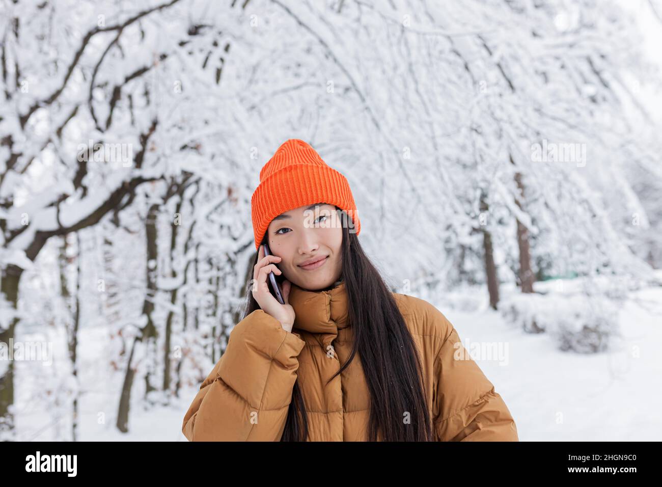 Eine schöne Chinesin in einem Winterpark telefoniert, warm gekleidet für einen Spaziergang Stockfoto