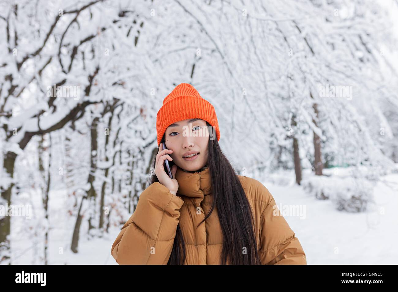 Eine schöne Chinesin in einem Winterpark telefoniert, warm gekleidet für einen Spaziergang Stockfoto