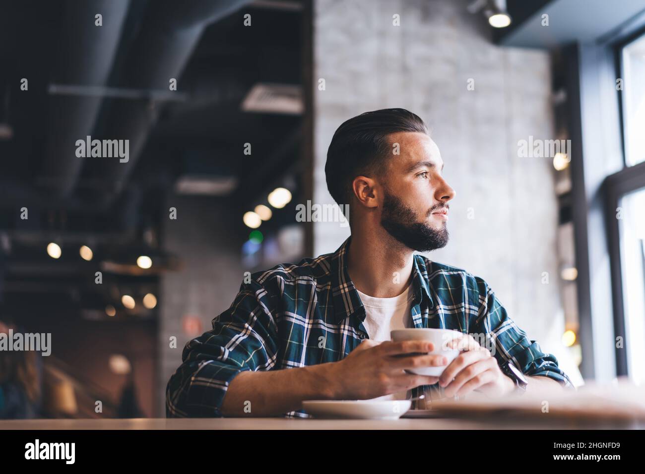 Nachdenklicher männlicher Kunde, der in trendigem Hemd gekleidet ist und während der Kaffeestunden in der Region wegschaut Stockfoto