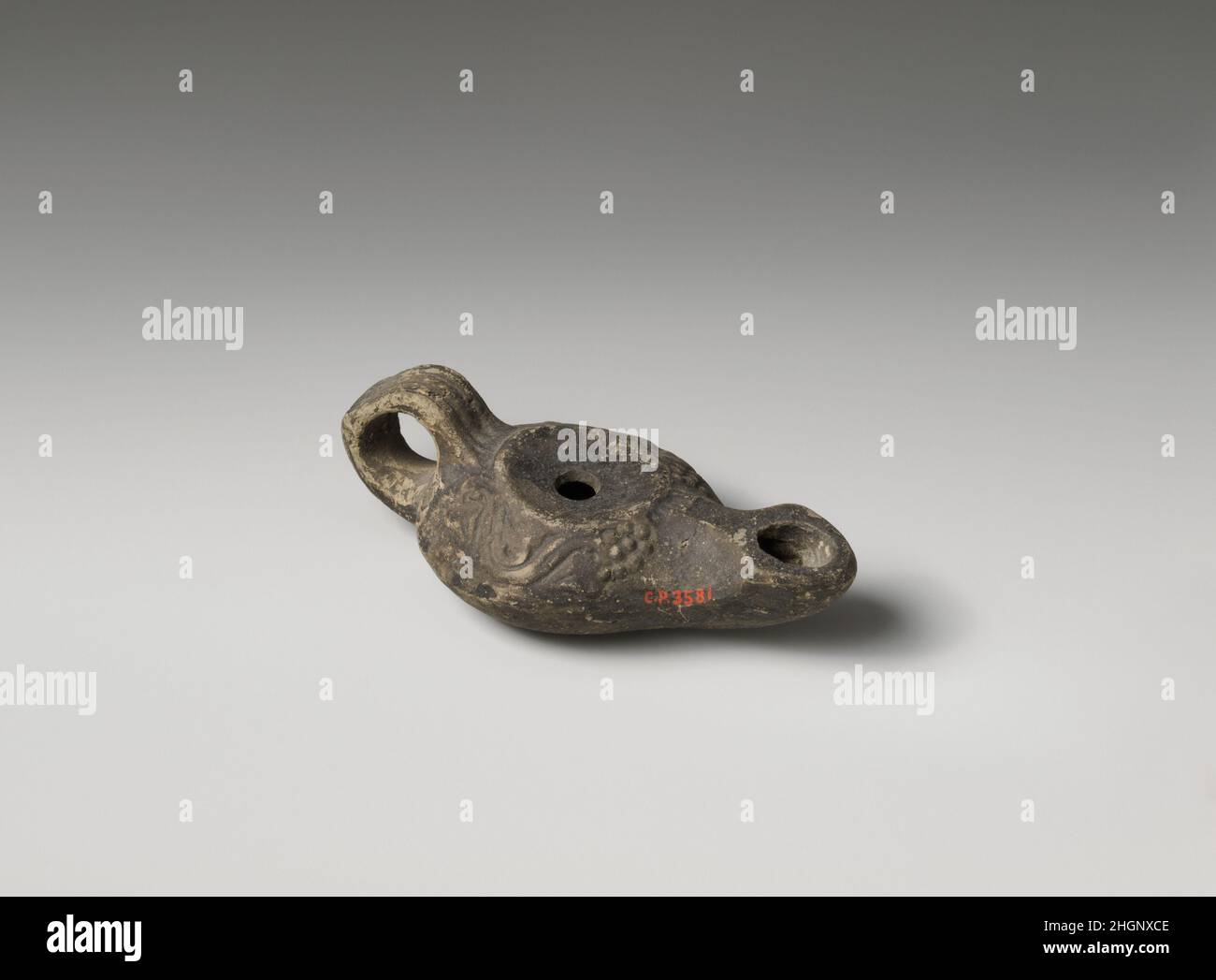Terrakotta-Öllampe aus dem 1st. Jahrhundert v. Chr., griechisch, mit Ringgriff. Einfarbige, konkave Diskus, mit zentraler Einfüllöffnung und angehobener Kante. Auf der konvex geschwungenen Schulter eine Rebe-Ranke, die vorne in ein Traubenbündel endet; eine kleine Franse ziert den Boden der Düse, die an den Seiten des großen Dochtlochs zeigt; auf dem Griff ein mittig von parallel eingeschnittenen Linien flankiertes Kabelmuster mit einem Querbalken auf der Rückseite; Hocke, karinierter Körper. Angehobener, breiter Basisring und flacher Boden.. Terrakotta-Öllampe. Griechisch. 1st Jahrhundert v. Chr.. Terrakotta. Hellenistisch. Terrakotta Stockfoto