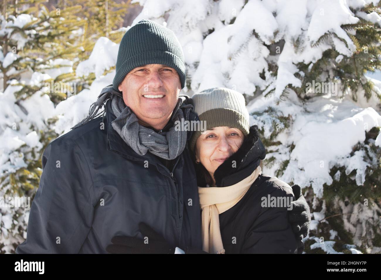 Ehepaar mittleren Alters in Winterkleidung umarmt sich gegenseitig und zeigt Zuneigung. Kaukasisches Paar lächelt zusammen zwischen Schnee und Wald. Candi Stockfoto