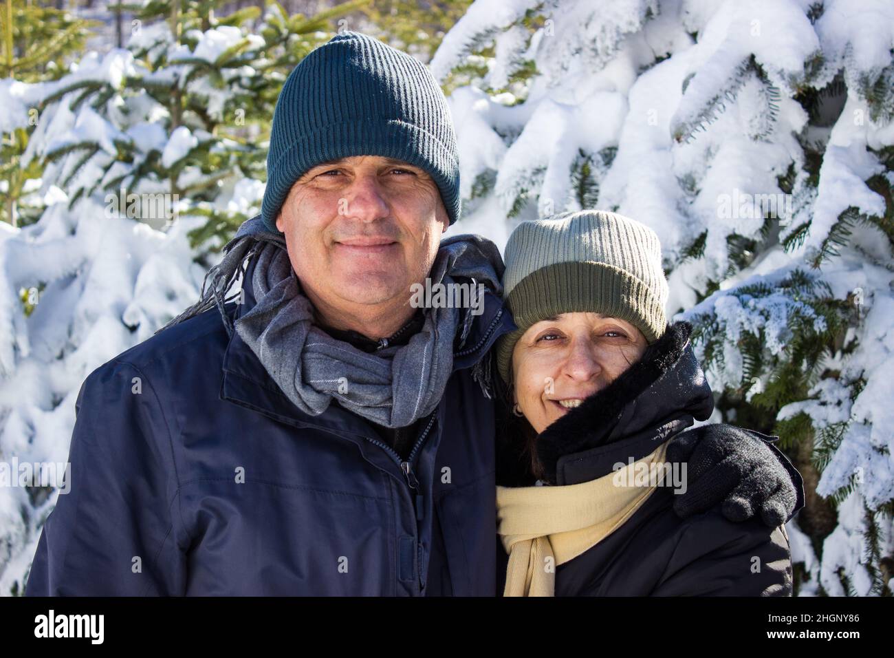 Ehepaar mittleren Alters in Winterkleidung umarmt sich gegenseitig und zeigt Zuneigung. Kaukasisches Paar lächelt zusammen zwischen Schnee und Wald. Candi Stockfoto