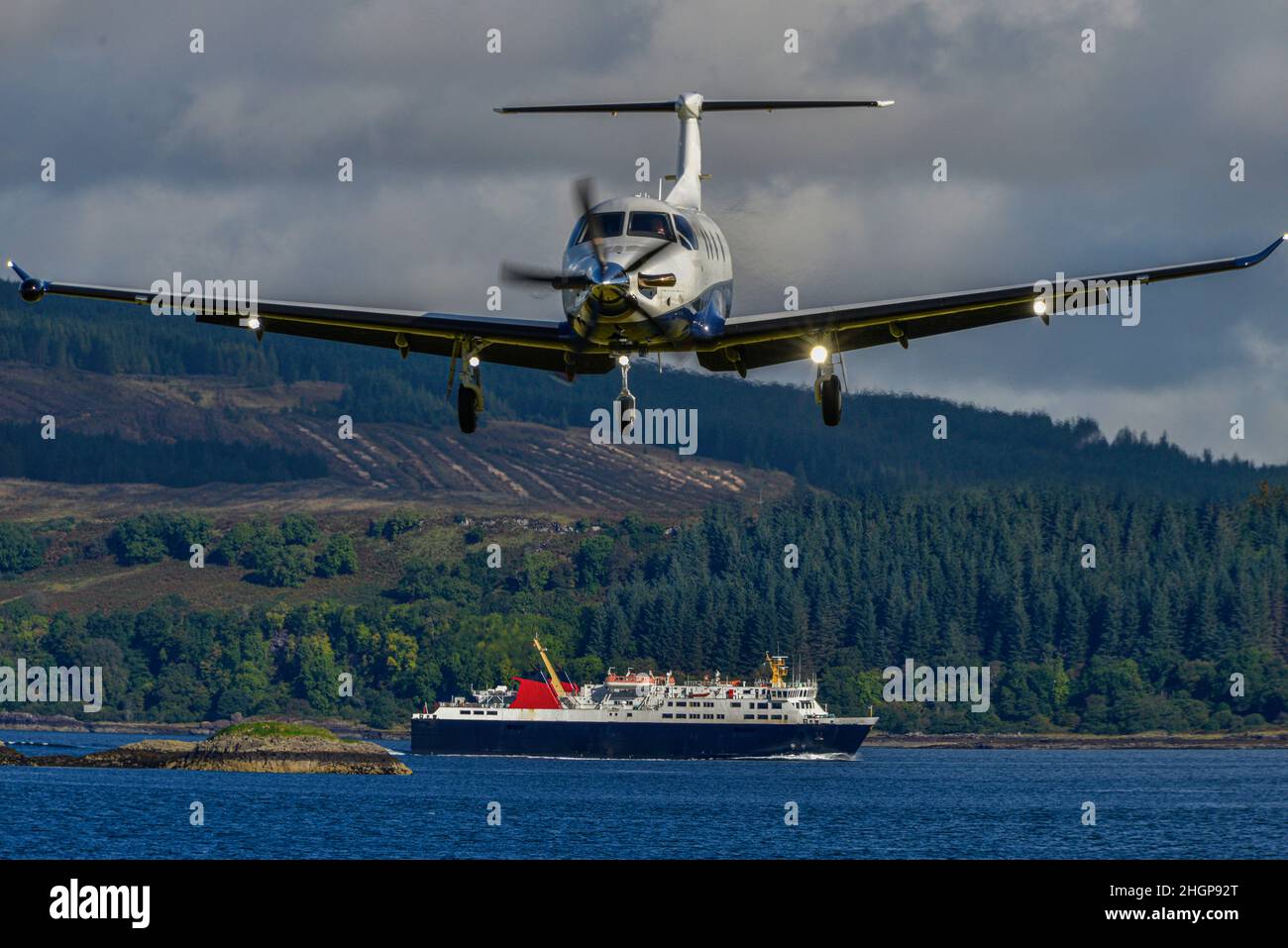 Ein Pilatus PC12-Flugzeug mit Unterwagen landet kurz vor der Landung. Im Hintergrund ist eine Inter-Island Fähre. Isle of Mull, Schottland. Stockfoto