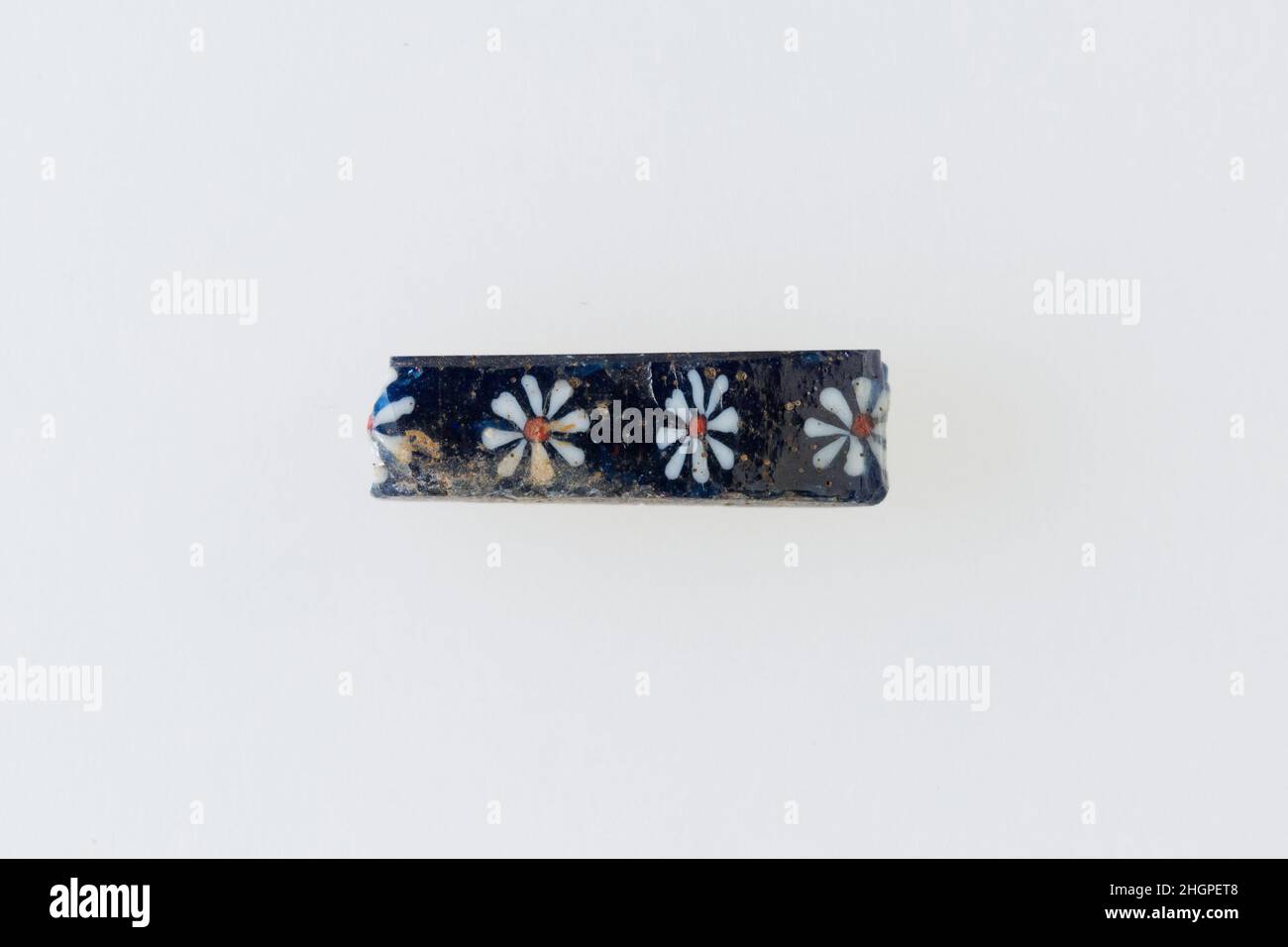 Inlay-Fragment mit Rosettenreihe 200 v. Chr.–100 n. Chr. Ptolemäische Periode–Römische Periode Eine Mosaikglas-Technik ermöglichte die Schaffung von Vielfachen eines Bildes: Eine figürliche oder gestalterische Komposition wurde durch Bündelung von farbigen Glasstöcken hergestellt, die dann zu einem langen Balken herausgezogen wurden. Die Stange wurde dann im rechten Winkel geschnitten, wahrscheinlich durch Aufschlagen der Stange mit geeigneten Werkzeugen, um kleine Inlay-Fliesen zu produzieren. Die Fliese würde dann geglättet und auf dem Gesicht poliert, das nach außen erscheinen soll.solche Elemente könnten auch nebeneinander verwendet werden, um sich wiederholende Muster zu erstellen. Inlay-Fragment mit Reihe von Rosetten. 200 V. CHR.– Stockfoto