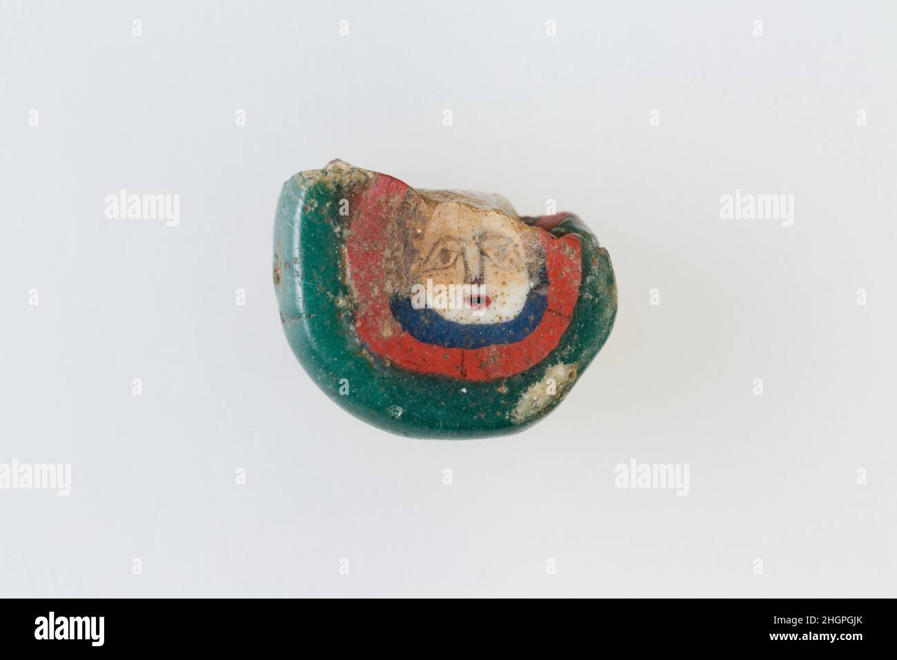 Inlay-/Perlenrohling mit Gesicht 100 v. Chr.–100 n. Chr. Ptolemäische Periode–Römische Zeit Eine Mosaikglas-Technik ermöglichte die Schaffung von Vielfachen eines Bildes: Eine figürliche oder gestalterische Komposition wurde durch Bündelung von farbigen Glasstöcken hergestellt, die dann zu einem langen Stab herausgezogen wurden. Die Stange wurde dann im rechten Winkel geschnitten, wahrscheinlich durch Aufschlagen der Stange mit geeigneten Werkzeugen, um kleine Inlay-Fliesen zu produzieren. Die Fliese würde dann geglättet und auf dem Gesicht poliert, das nach außen erscheinen soll. Inlay-/Perlenrohling mit Stirnseite. 100 V. CHR.–100 N. CHR. Glas. Ptolemäische Periode–Römische Periode. Aus Ägypten Stockfoto