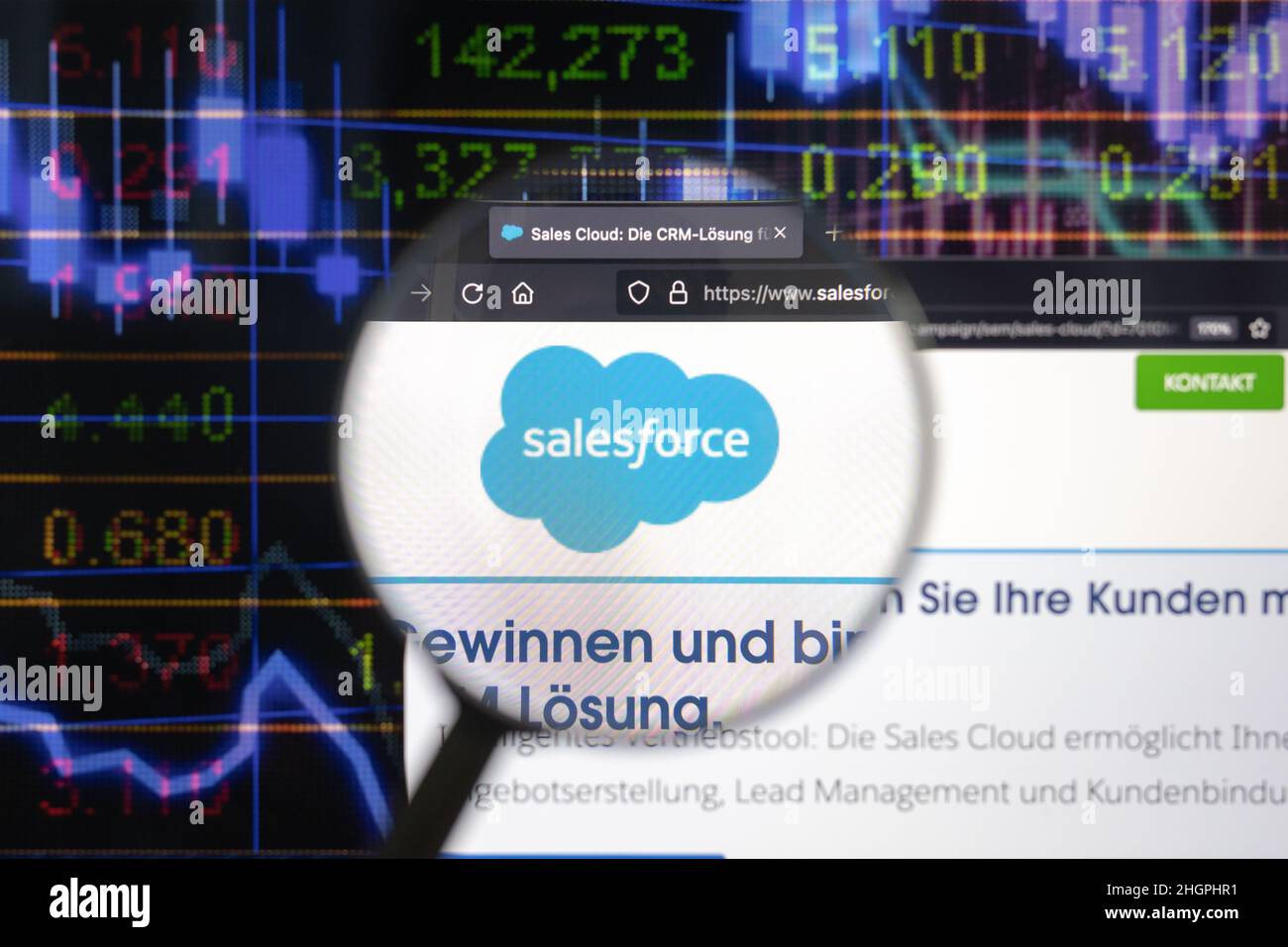 Salesforce-Firmenlogo auf einer Website mit verschwommenen Börsenentwicklungen im Hintergrund, die auf einem Computerbildschirm durch eine Lupe gesehen werden. Stockfoto