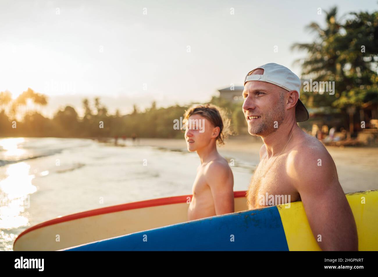 Lächelnder Mann mit jungendlichen Jungen Sohn mit Surfbrettern geht zum Surfen ins Meer. Sie haben einen Winterurlaub und genießen ein wunderschönes Sonnenuntergangslicht. Familie Stockfoto