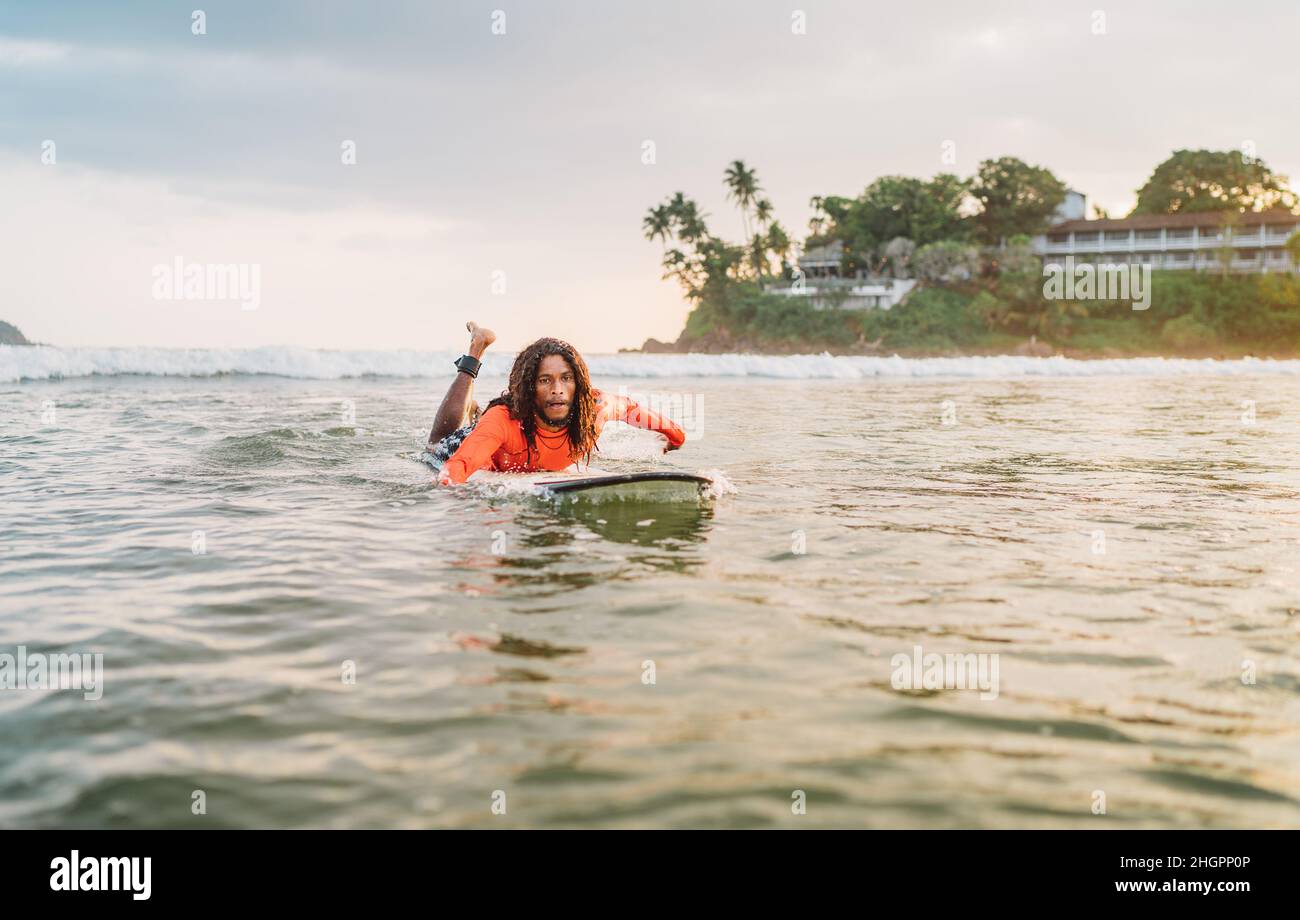Schwarzer langhaariger Mann, der auf einem langen Surfbrett zum Surfspot im Indischen Ozean paddelt. Palmenhain übersäte Sonnenstrahlen im Hintergrund. Extreme Wasser e Stockfoto