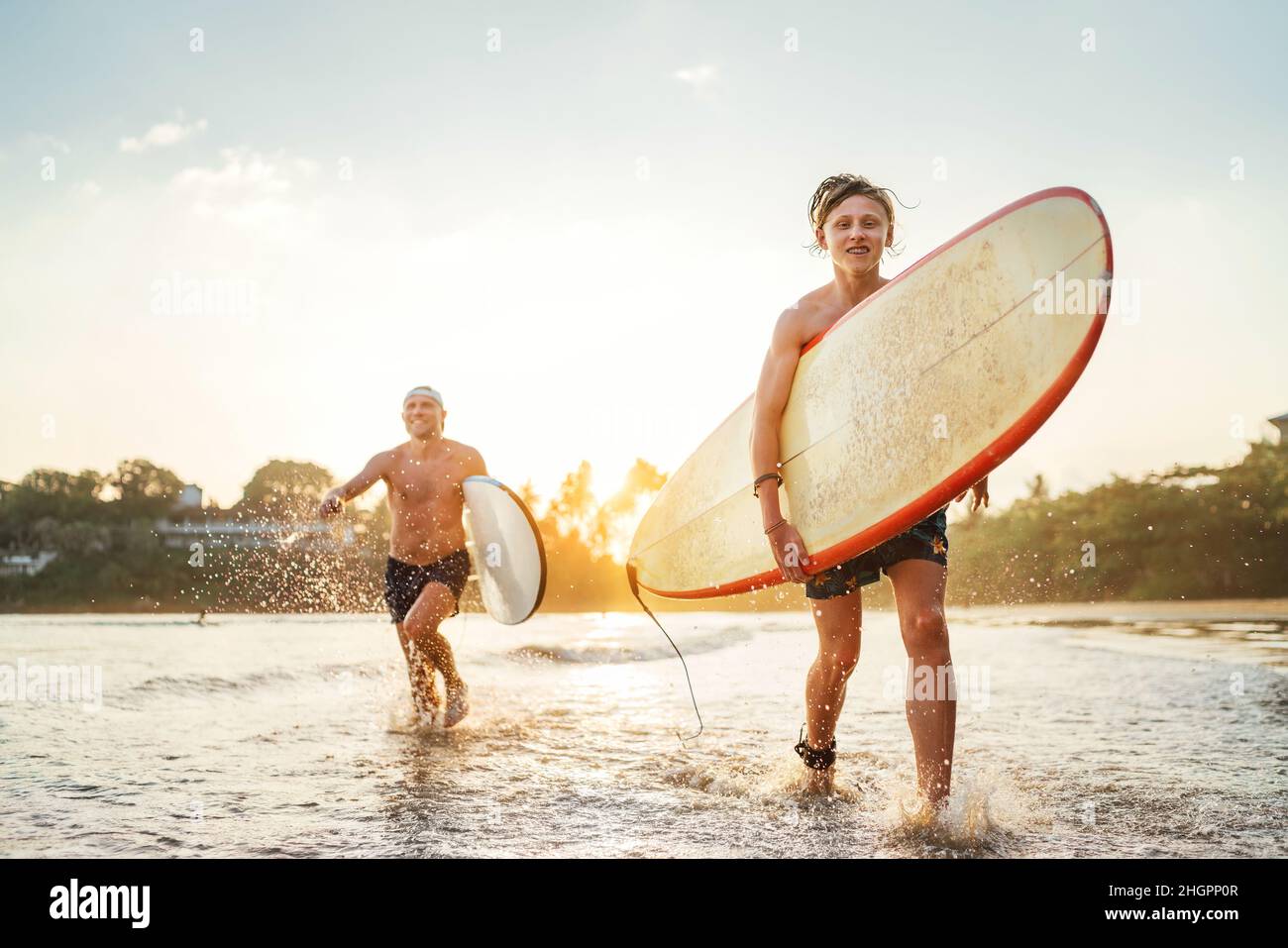 Junger Teenager mit einem Surfbrett, der nach dem Surfen mit seinem Vater am Sandstrand des Ozeans läuft. Sie lächeln und genießen ein wunderschönes Sonnenuntergangslicht. Fa Stockfoto