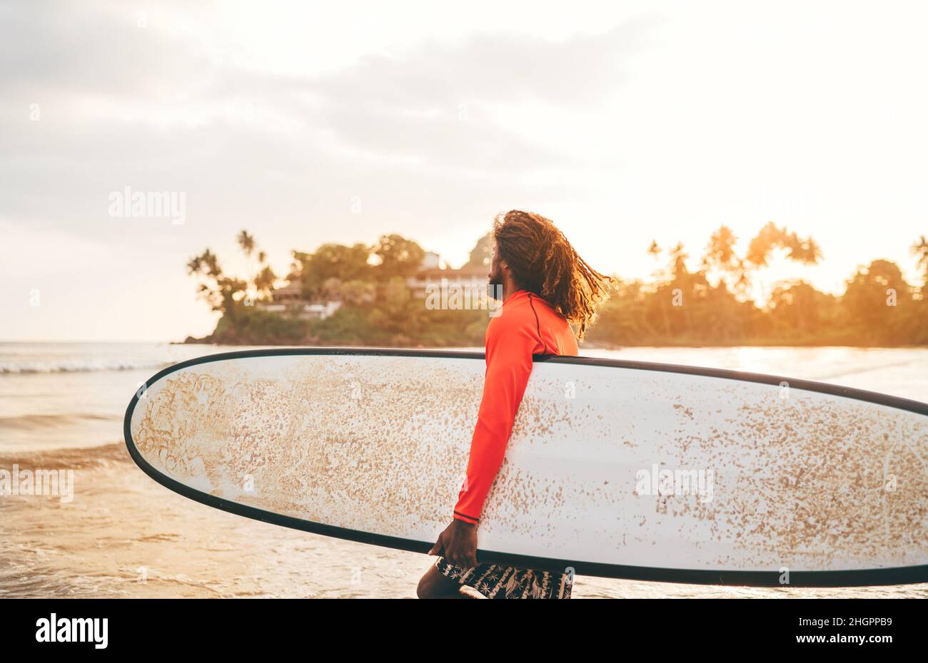 Schwarzer langhaariger Teenager-Junge mit einem gewachsten Surfbrett, bereit zum Surfen mit Sonnenuntergang-Hintergrundbeleuchtung. Er läuft in die Wellen des indischen Ozeans. Extremwassersport und Stockfoto