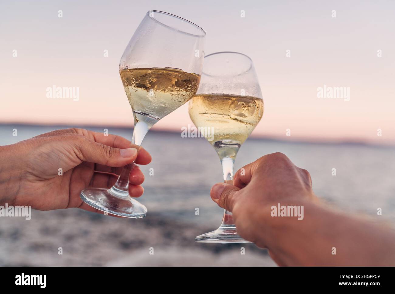 Verliebte Paare klirren mit Weißweingläsern, während sie den wunderschönen rosa Sonnenuntergang am Meer genießen. Romantischer Urlaub oder Dating-Konzept aus nächster Nähe im Stockfoto