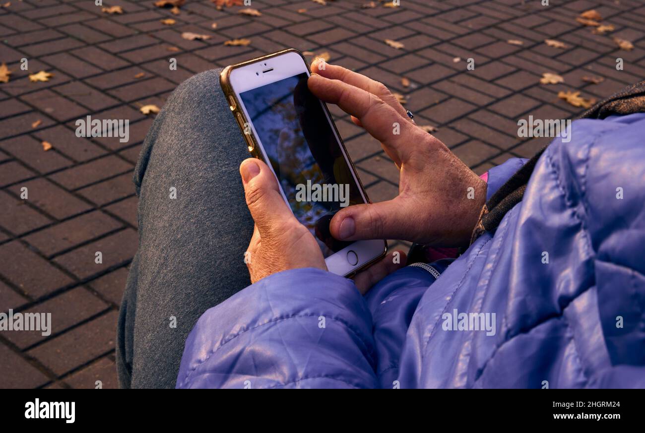 Eine Frau hält ein Mobiltelefon in den Händen vor dem Hintergrund herbstlicher Blätter, die auf dem Bürgersteig verstreut sind. Stockfoto