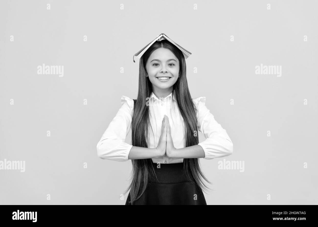 Glückliches Teenager-Mädchen in Schuluniform mit Buch auf Kopf meditieren mit Handbewegung, Ruhe bewahren Stockfoto
