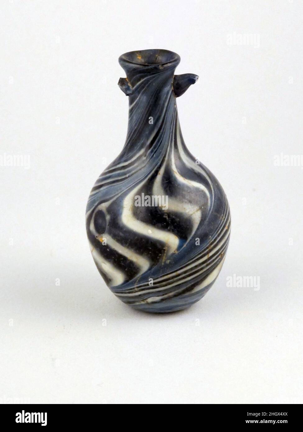 Parfümflasche aus Glasmosaik Anfang des 1st. Jahrhunderts n. Chr. römisch Durchscheinend Kobaltblau, mit marmorierten Spuren in opak-weiß; Griffe in gleichen Farben. Abfackeln, abgerundeter Rand; Hals, der sich nach unten bis zur schrägen Schulter ausdehnt; Knollenkörper mit nach unten verjüngenden Seiten; kleiner konkaver Boden; Zwei Griffe oben am Hals angebracht und nach außen gezogen, ohne Spuren von Befestigung an Schulter oder Körper.Trails bedecken das gesamte Schiff mit dicken Wirbeln auf der Unterseite und am Körper Bänder aus feinen parallelen Linien, die sich mit dicken S-förmigen Trails abwechseln; Auf einem der S-förmigen Trails erscheint ein großer blauer Punkt, der unten und unten geknackt ist Stockfoto