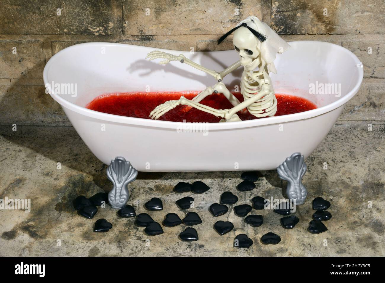 Anti-Valentinstag-Skelett in der Badewanne gefüllt mit blutfarbener Himbeer-Gelatine und schwarzen Herzen auf dem Boden verstreut Stockfoto