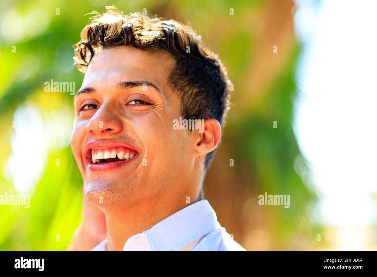 Hübscher Mann in weißem Hemd mit einem strahlend weißen Lächeln und Blick auf die Kamera im Sommerpark Stockfoto