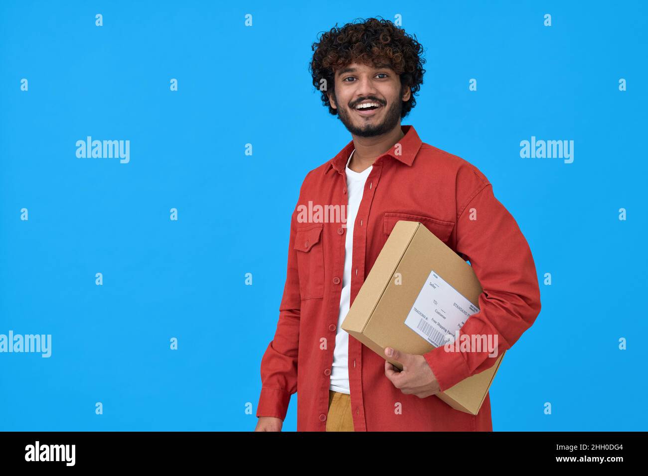 Glücklicher indischer junger Mann, der Paketkasten auf blauem Hintergrund isoliert hält. Stockfoto