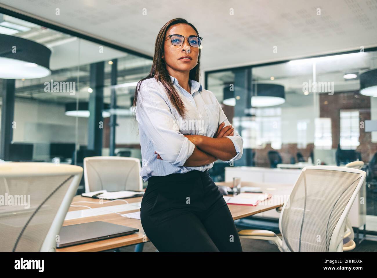 Selbstbewusste Geschäftsfrau, die in einem modernen Büro nachdenklich aussieht. Eine Geschäftsfrau mittleren Erwachsenen, die sich mit gekreuzten Armen auf einen Tisch lehnte. Unternehmerin w Stockfoto