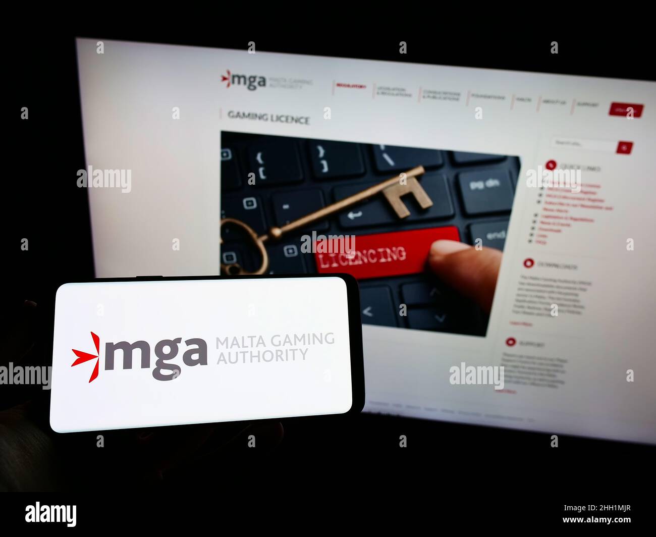 Person, die Mobiltelefon mit dem Logo der maltesischen Institution Malta Gaming Authority (MGA) auf dem Bildschirm vor der Webseite hält. Konzentrieren Sie sich auf die Telefonanzeige. Stockfoto