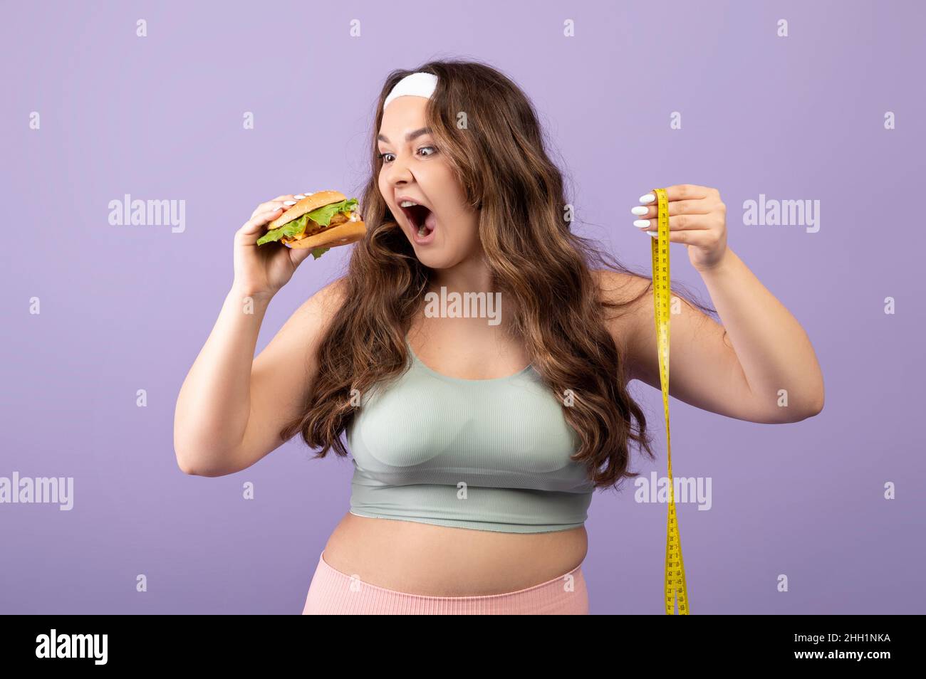 Lustige hungrige kaukasische plus Größe junge Frau in Sportkleidung mit Maßband isst appetitlichen Burger Stockfoto