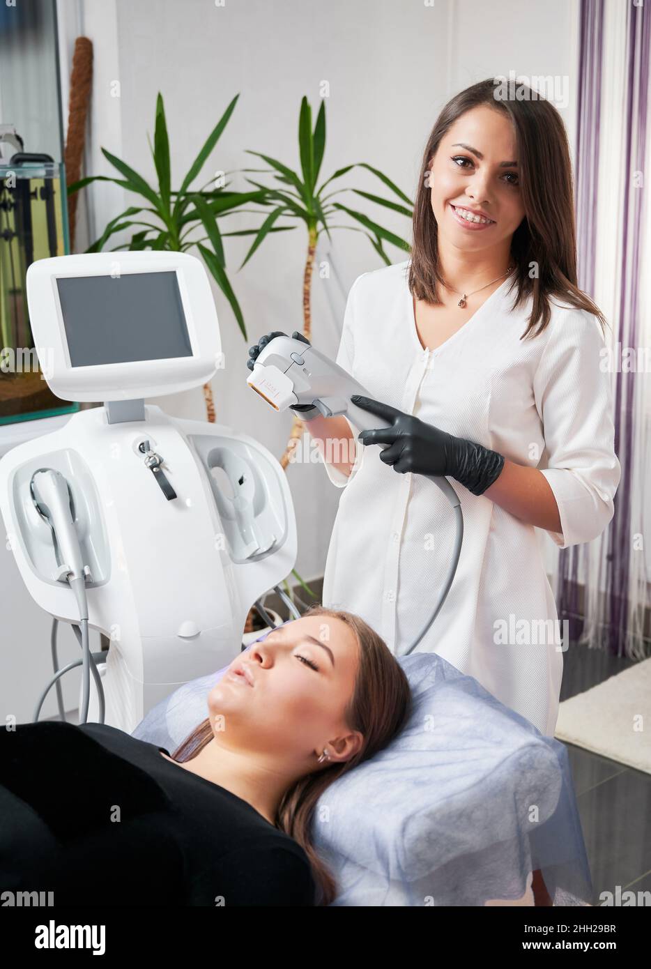 Fröhliche junge Frau Kosmetologin, die ein medizinisches Ultraschallgerät hält und lächelt, während sie beim Kunden steht. Charmante weibliche Patientin, die auf dem Tagesbett liegt, während sie in einer modernen Klinik ein SMAS-Hebeverfahren durchgeführt hat. Stockfoto