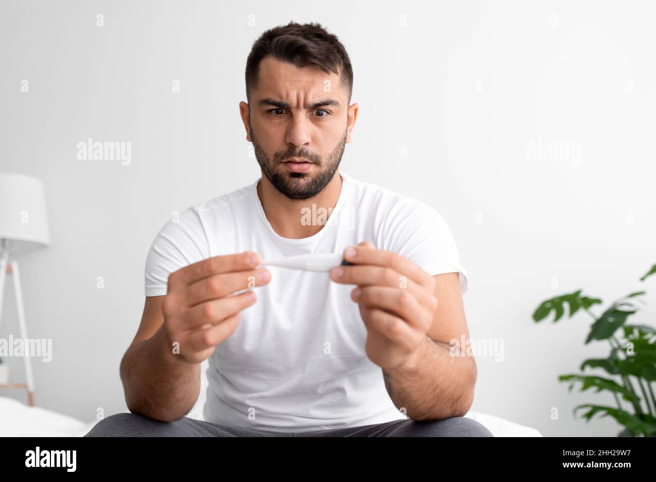 Traurig schockiert jungen Mann mit Bart schaut Thermometer mit hoher Temperatur und leidet an krank allein Stockfoto