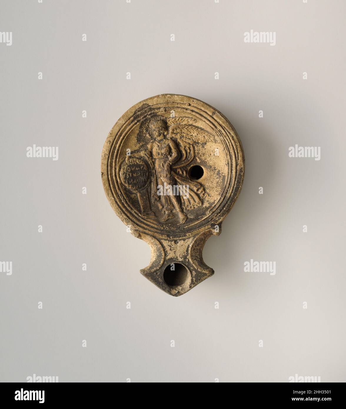 Terrakotta-Öllampe A.D. 1–50 Roman Loeschcke Typ 1A. Hergestellt. Discus: In Hochrelief, geflügelter Nike (Victory) stehend, nach links zeigend, mit eingeschriebenem Schild über dem Altar; ein einzelnes Feilloch in der Mitte rechts hinter Nike, mit einem Band aus Linien und Rillen zum Rand hin. Spiraldüsen flankieren die Winkeldüse. In einem erhöhten Basisring, umrandet durch zwei eingeschnittene Kreise, einer flachen Basis, mit eingeschnittenen Buchstaben in der Mitte: FAVSII.auf dem Schild in Latein ist ein Neujahrswunsch nach Glück und Glück für den Besitzer: ANNV / NOVM FAV/STVM FEL/ICEM MI/HI.intakt.auf Diskus, geflügelter Sieg mit eingeschriebenem Schild. Ein Stockfoto