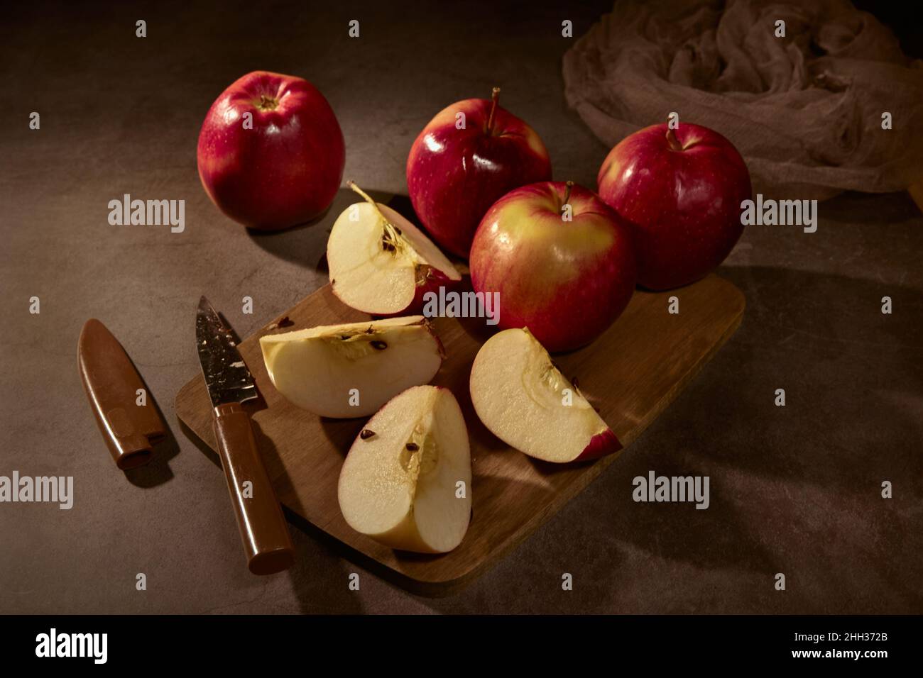Apfelbrett. Äpfel und ein Messer stehen auf einem Schneidebrett, ein Apfel wird in vier Stücke geschnitten und die ganze Komposition auf dunklem Hintergrund Stockfoto