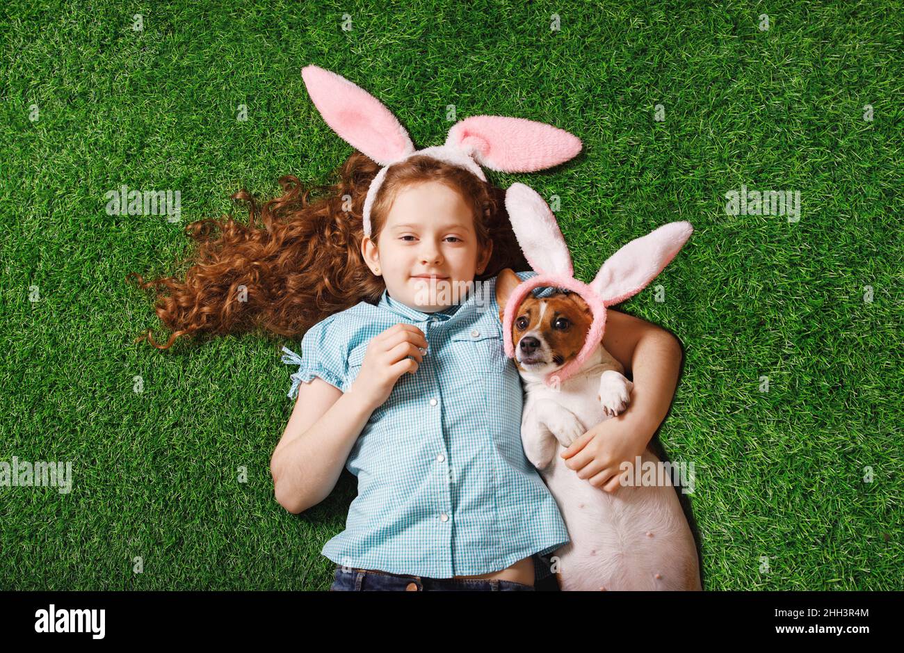 Nettes lockiges Mädchen mit fetten Hund mit Kaninchenohren auf grünem Gras liegen. Osterkonzept. Stockfoto