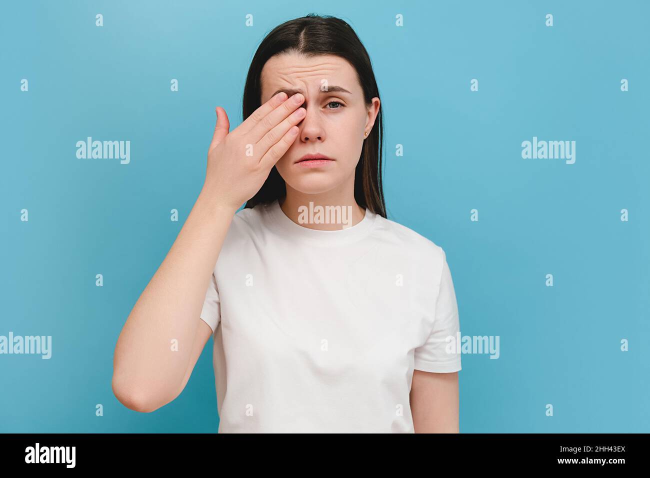 Junges Mädchen, das unter Augenschmerzen leidet und etwas in den Augen fühlt, posiert an der blauen Wand. Ursache von Schmerzen sind Kontaktlinsenproblem, Konjunktivitis, für Stockfoto