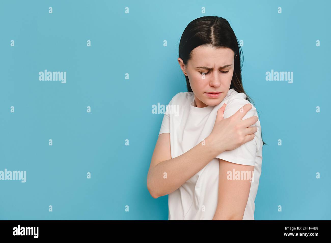 Unglücklich traurig müde junge Brünette Frau hat schmerzhafte Gefühle in der linken Schulter, kleckert Zähne, trägt weißes T-Shirt, posiert isoliert auf blauem Studiorücken Stockfoto