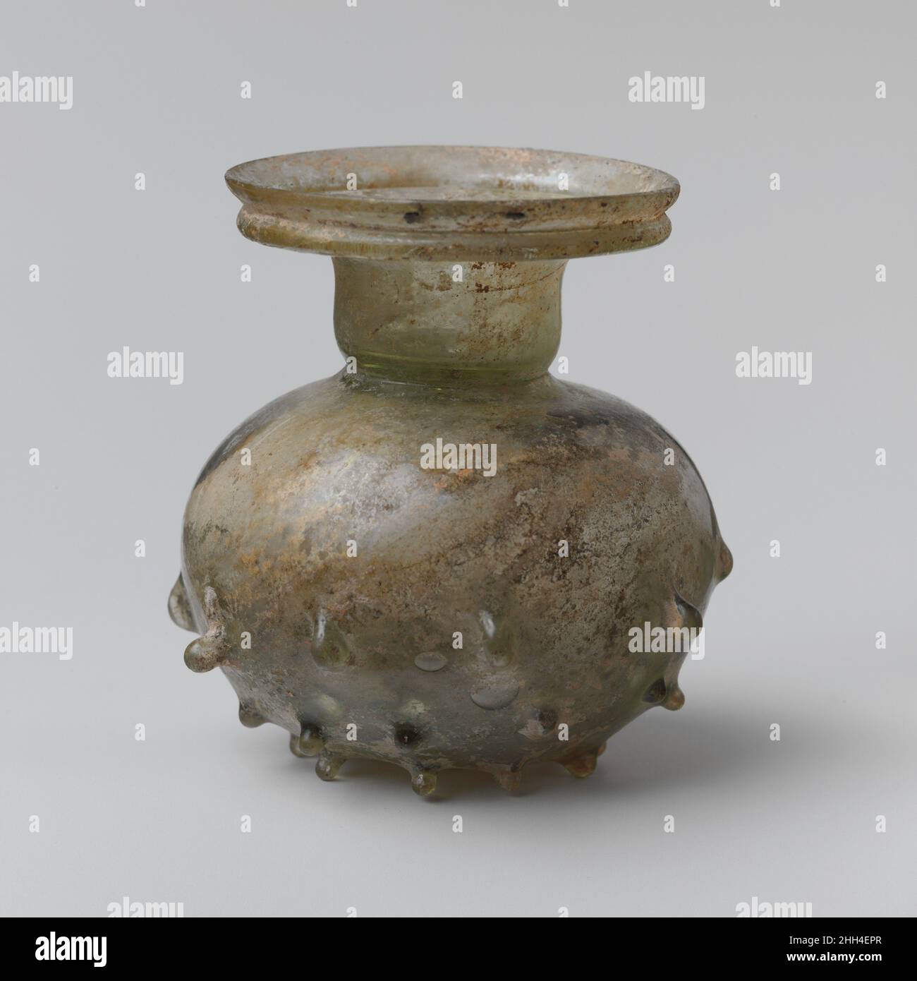 Glas-Sprinklerkolben 3rd–4th Jahrhundert n. Chr. römisch, östliches Mittelmeer Durchscheinend hellgrün. Ungleichmäßiger, abgerundeter Rand, nach oben abfackend, mit hervorstehendem gefalteten Flansch unten; breiter Mund; kurzer zylindrischer Hals, leicht an der Basis gebogen, mit gefaltetem Diaphragma mit kleinem Loch in der Mitte; Kugelkörper; Flacher Boden, in der Mitte leicht eingedrückt, mit kleiner runder Pontil-Markierung.auf der unteren Körperhälfte drei unregelmäßige horizontale Reihen von verschraubten Knöpfen (11 in der oberen Reihe, 12 in der Mitte und 9 in der unteren Reihe).intakt; ein paar Blasen und schwarze Verunreinigungen; etwas kalkige Bodenverkrustungen, cremige Verwitterung und iride Stockfoto