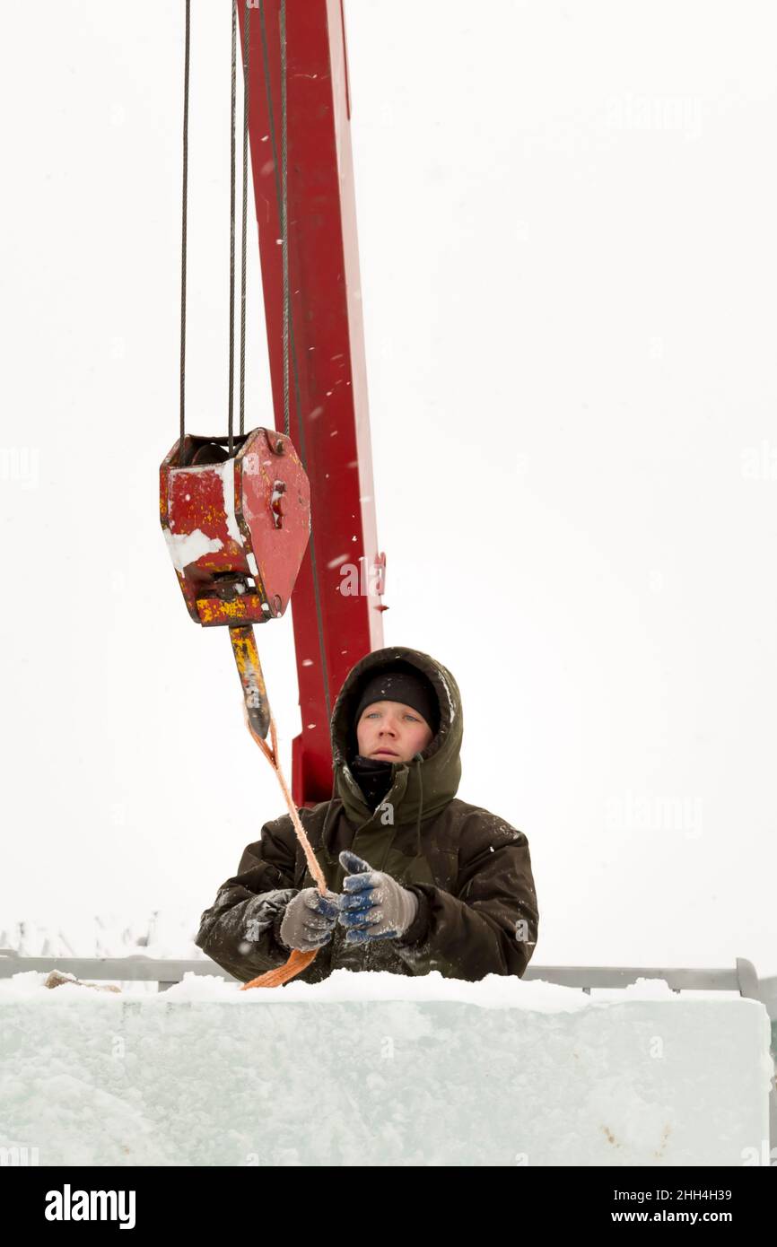 Porträt eines Arbeiters in einer grünen Jacke mit einer Kapuze am Haken eines LKW-Krans, der Eisblöcke lädt Stockfoto