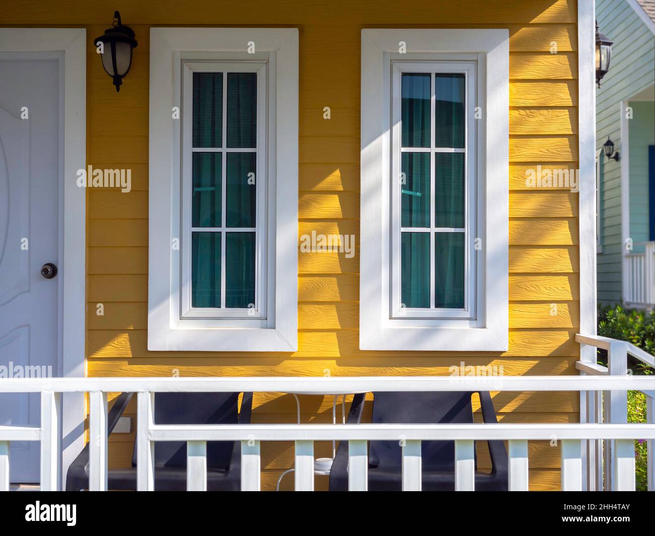 Zwei weiße Hausfenster mit Glas, grüner Vorhang Dekoration auf dem gelben Holz Bewohner in der Nähe der Haustür und Terrasse, Blick von draußen. Stockfoto