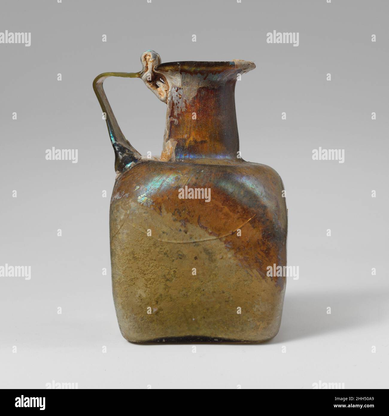 Quadratische Glasflasche 2nd–3rd Jahrhundert n. Chr. Römische kleine quadratische eingriffige FlaschenDurchscheinend hellgelb-grün; Griff in gleicher Farbe.einfacher Rand mit abgeschrägter Lippe bis zum abfackelnden Mund; zylindrischer Hals, der sich nach unten ausdehnt; eingedrückte, horizontale Schulter mit abgerundeten Ecken; vier flache Seiten, nach unten zulaufend; leicht konkaver Boden mit leichten Rippen in parallelen Linien; Spuren von großen kreisförmigen Pontil-Markierung; Griff als breites Pad entlang der äußeren Kante des Sholuders angebracht, gezogen und nach außen als schmale Gurt, dann horizontal gedreht und an der Spitze des Halses und der äußeren Kante des Randes mit Schlaufe über r geschleppt Stockfoto