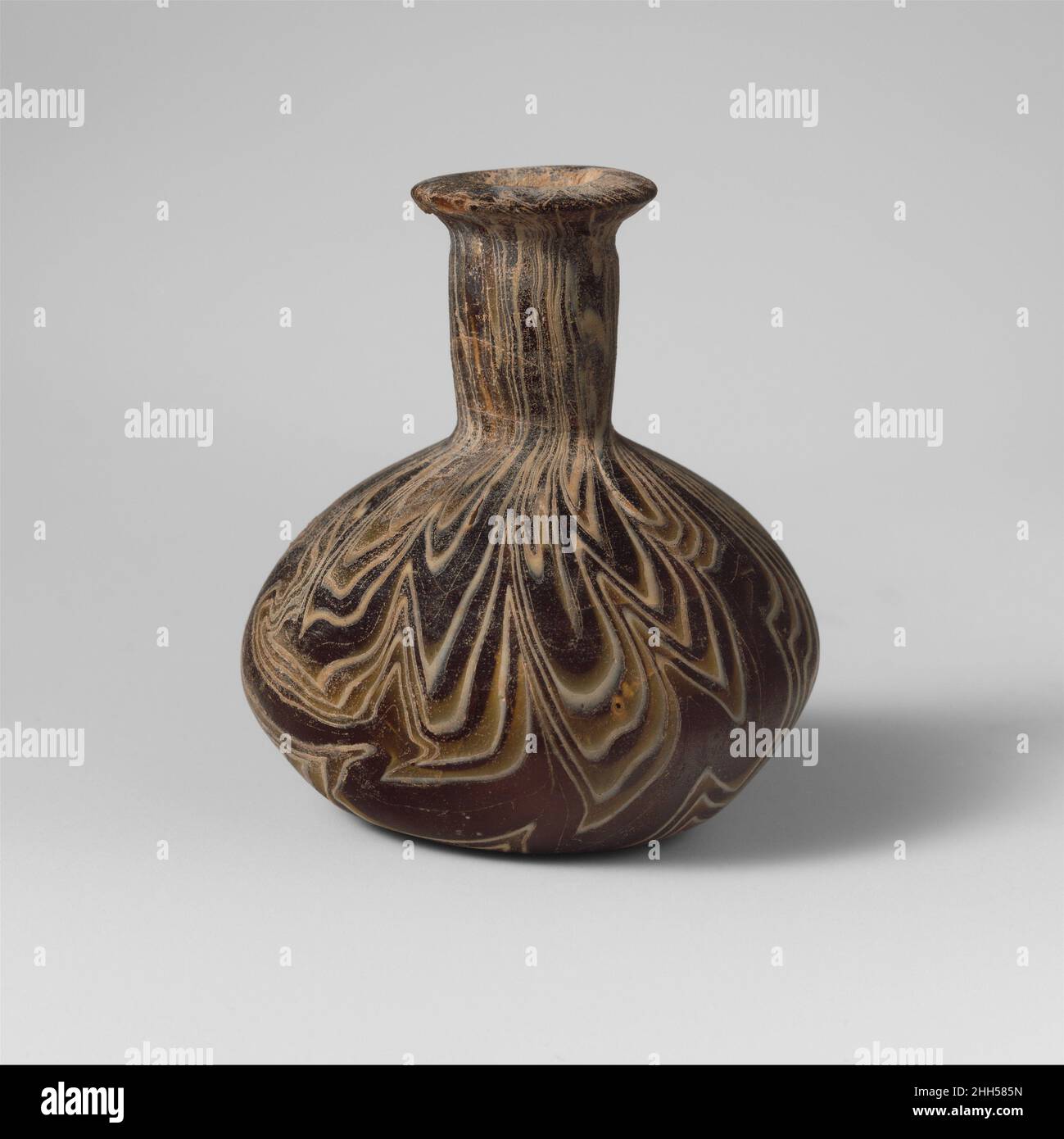 Glasmosaikflasche 1st, Hälfte des 1st. Jahrhunderts n. Chr., römisch, lichtdurchlässig, honigbraun und opak-weiß, leicht abgeschrägter Rand mit kantiger Außenlippe; zylindrischer Hals, nach unten zugeneigt, mit Werkzeugeinbuchtung um den oberen Rand unter dem Rand; hockiger Kugelkörper; Leicht konkaver Boden.Marmorierte Mosaikmuster aus Abschnitten eines einzelnen Rohres in einem braunen Boden mit weißen parallelen Linien in einem unregelmäßigen Zickzack-Muster gebildet.intakt; stumpfend, leichte Lochfraß, weißlich irisierende Verwitterung, und innen Hals und Körper Bodenverkrustungen.Rotary Schleifspuren auf der Außenseite.. Glasmosaikflasche. Roman. 1st Hälfte von 1st Cen Stockfoto