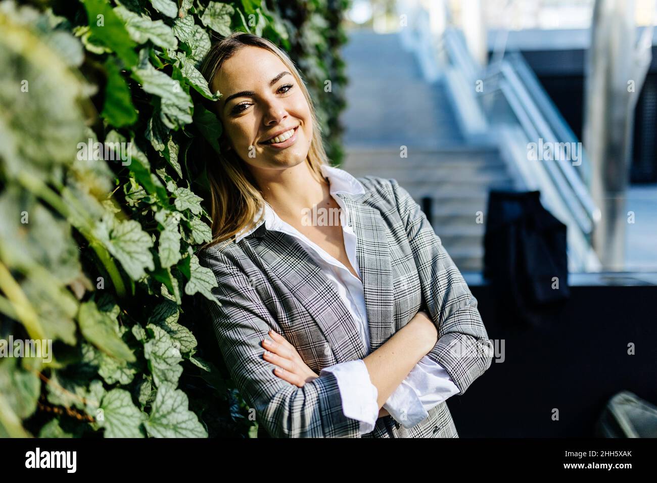 Lächelnde Geschäftsfrau mit gekreuzten Armen, die sich auf grünen Efeu-Pflanzen stützt Stockfoto