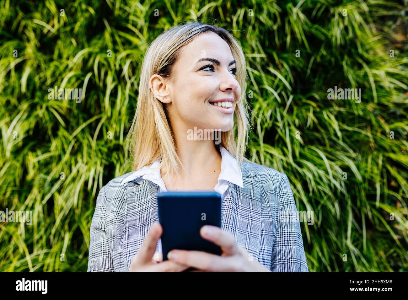 Junge blonde arbeitende Frau mit Smartphone vor grünen Pflanzen Stockfoto