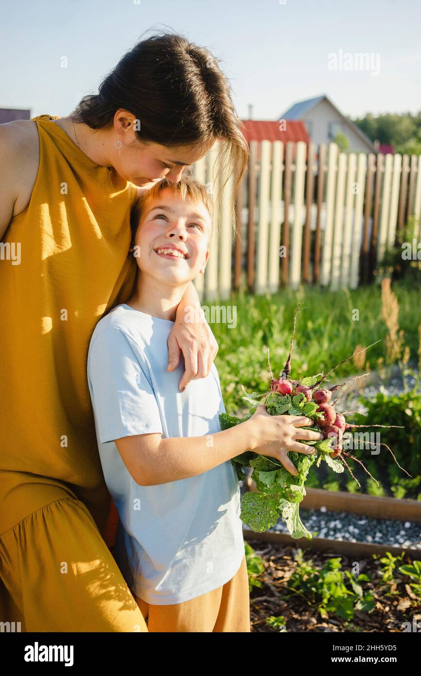 Glückliche Mutter umarmt lächelnden Sohn hält Rettich im Garten Stockfoto