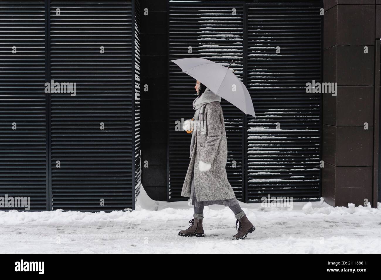 Kontemplative Frau mit Regenschirm, die im Winter auf einem Wanderweg an einer Wand spazieren geht Stockfoto