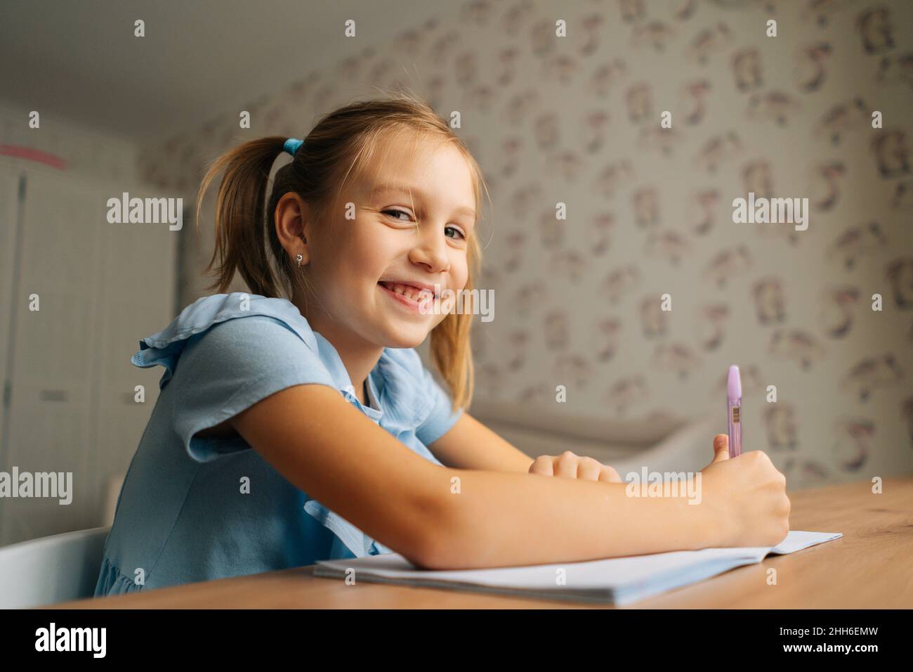 Nahaufnahme von lächelndem, elementarem kleinen Mädchen, das Hausaufgaben macht und am heimischen Tisch am Fenster sitzt und die Kamera anschaut. Stockfoto